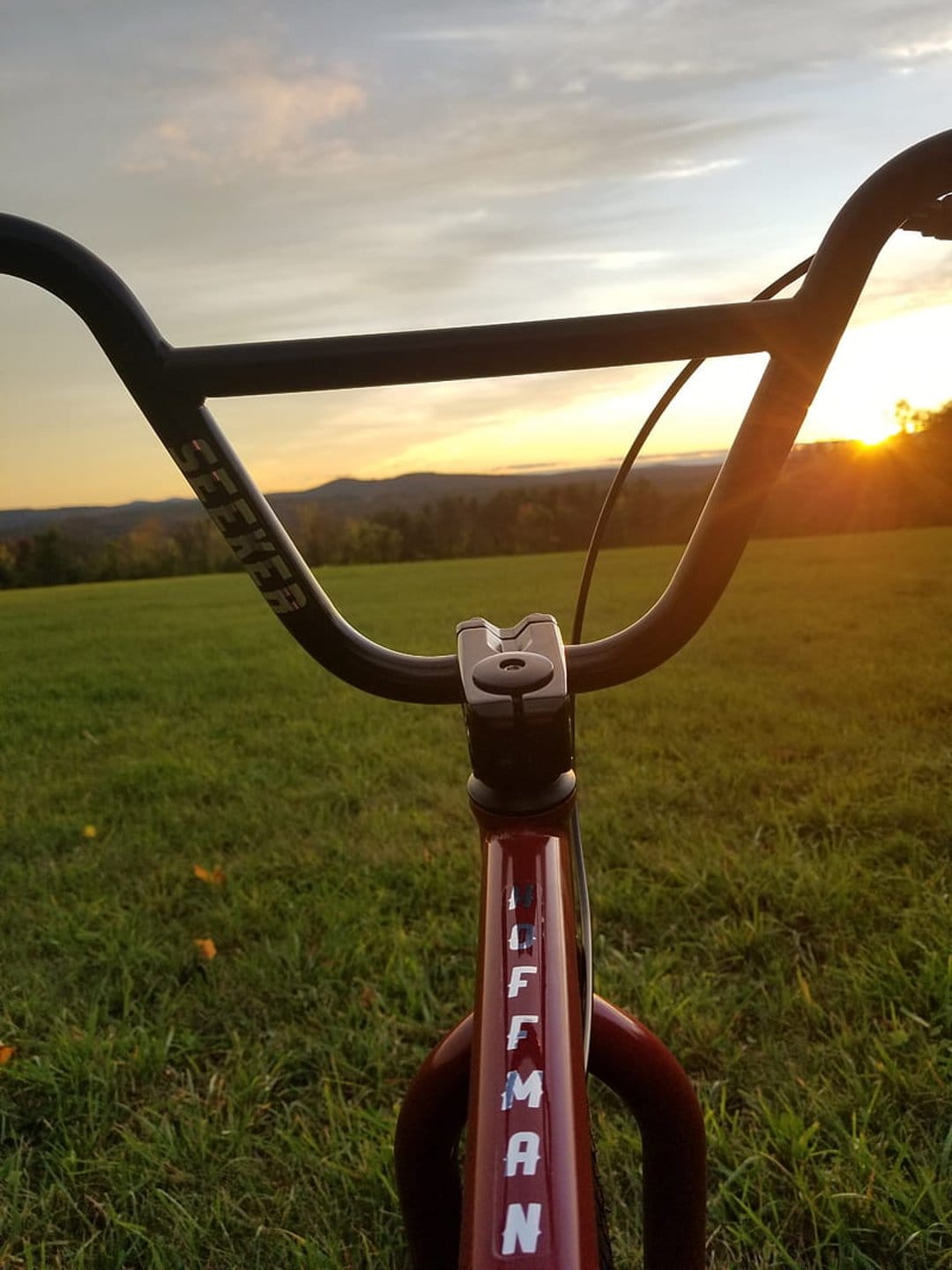 BMX handlebars with Seeker and Hoffman branding overlook a grassy field and hills during a golden sunset.