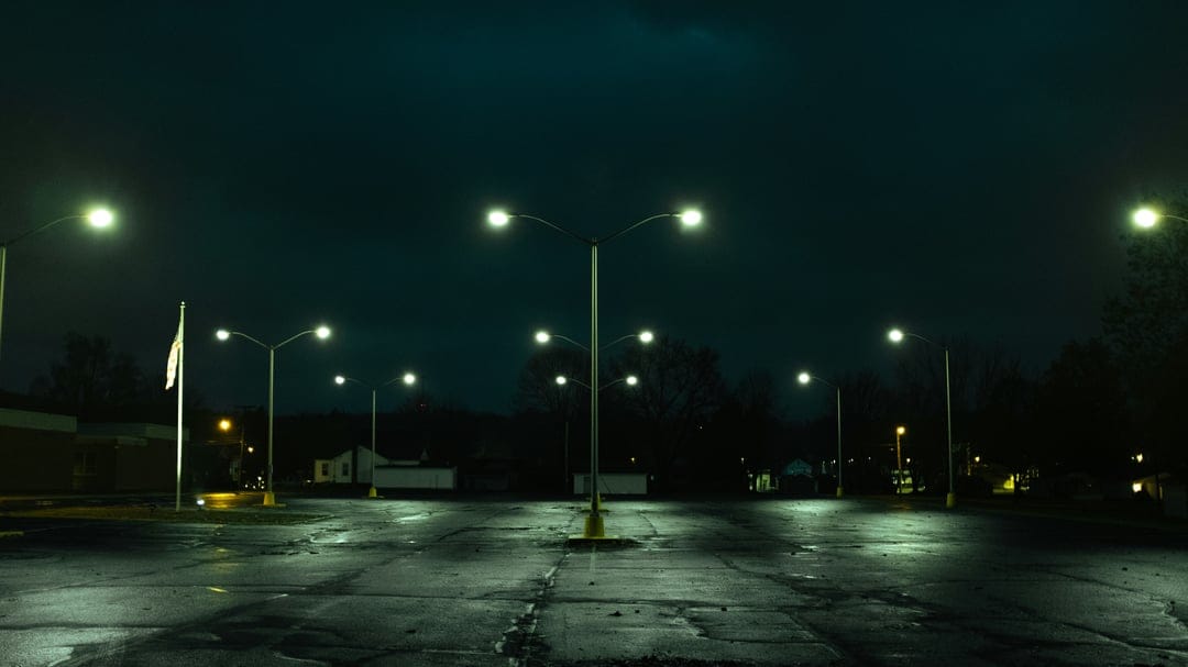 Empty asphalt parking lot at night with glowing streetlights casting long shadows on wet, cracked pavement.