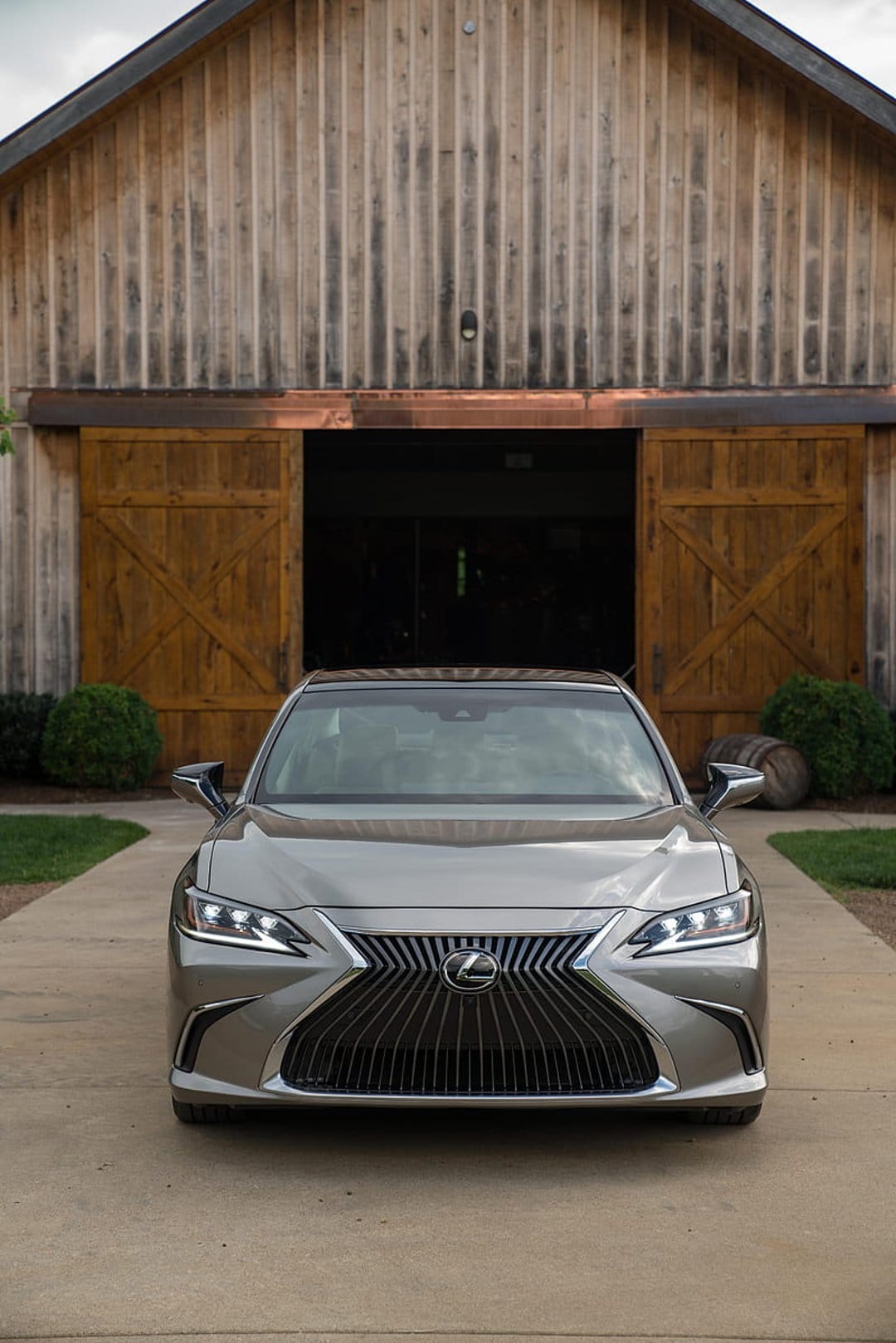 Silver Lexus sedan with glowing headlights parked in front of a rustic wooden barn on a concrete driveway.
