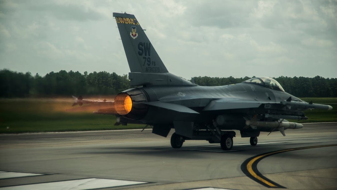 F-16 fighter jet with a bright orange afterburner flame on a tarmac under an overcast sky during takeoff.