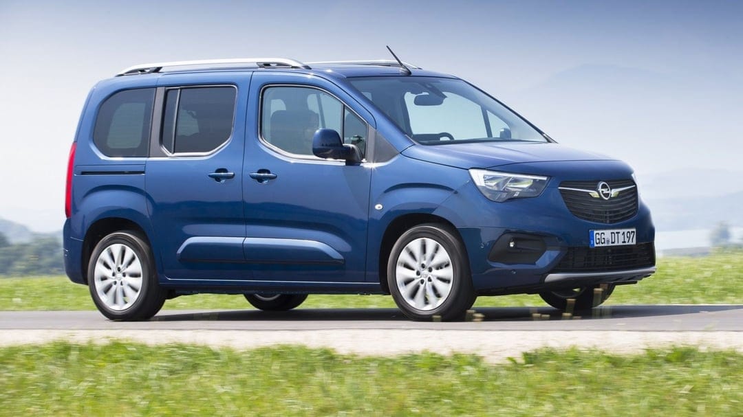 Blue Opel Combo Life van driving on a road past green fields under a clear sky with motion-blurred wheels.