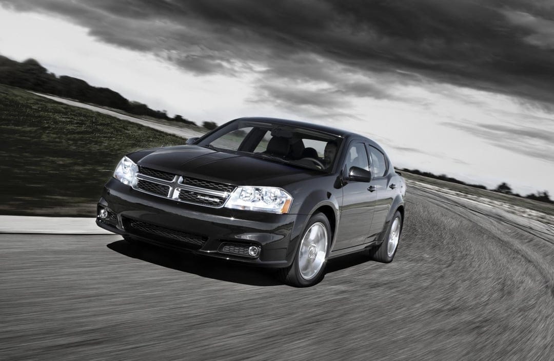 Black Dodge Avenger sedan driving fast on a curved asphalt track under a moody, dramatic cloudy sky.