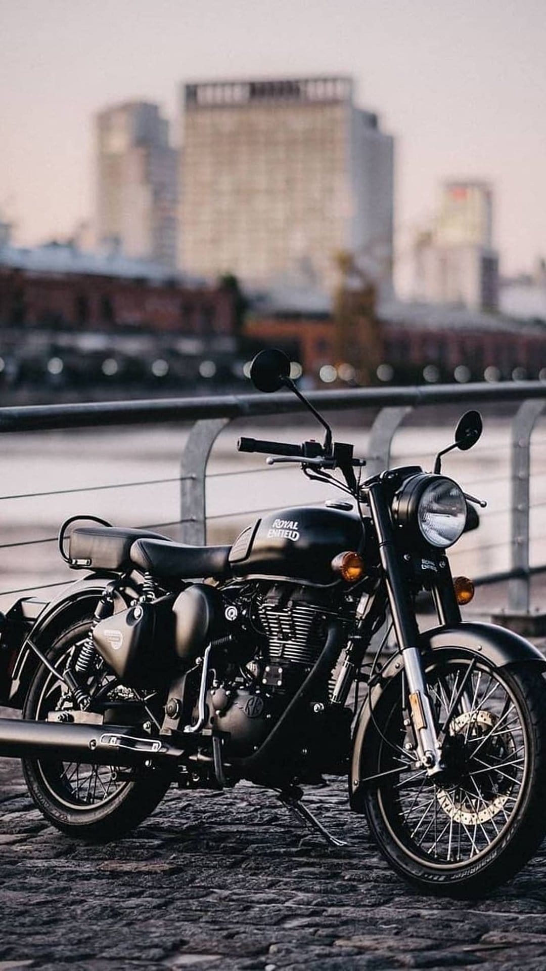 Black Royal Enfield motorcycle on a cobblestone street by a lake at sunset with blurred city buildings behind.