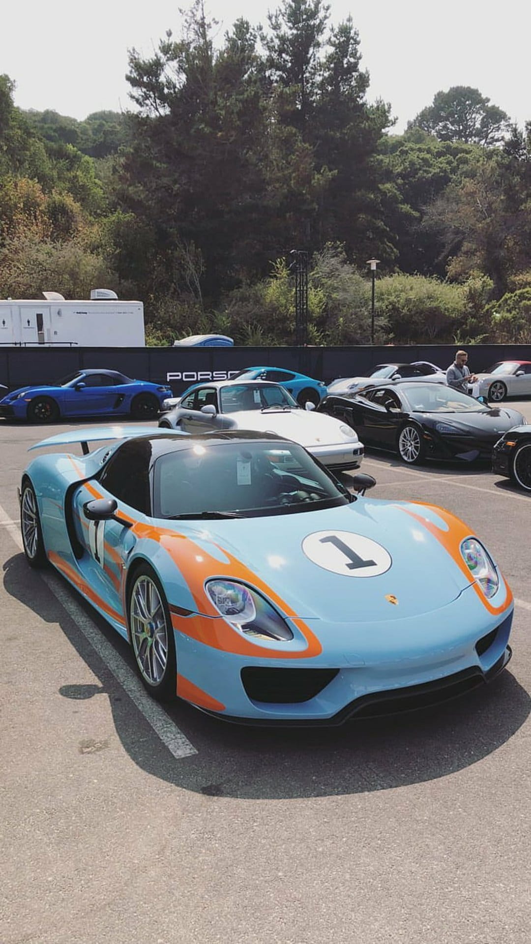 Porsche 918 Spyder in light blue Gulf livery with orange stripes parked outdoors at an automotive exhibition.