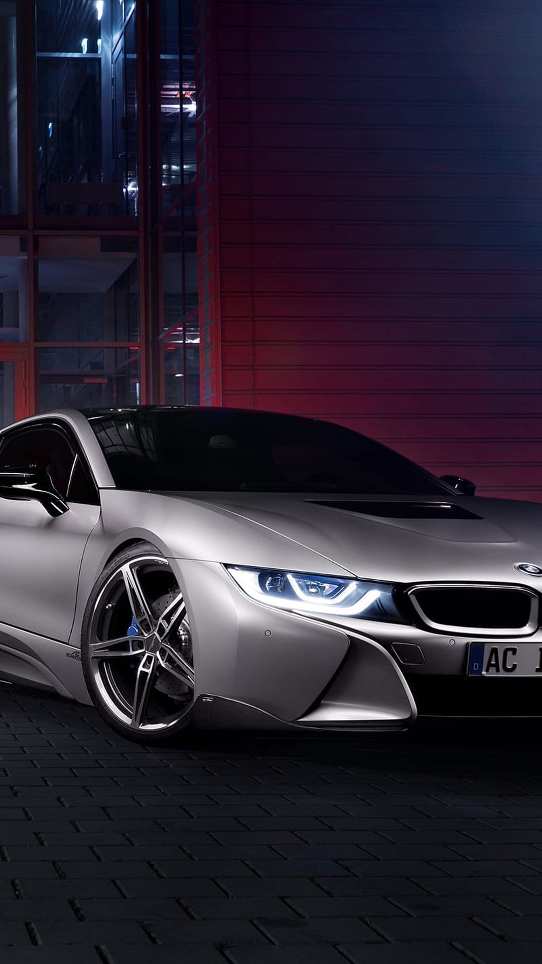 Silver BMW i8 sports car with blue glowing headlights parked on a brick surface near a red wall at night.