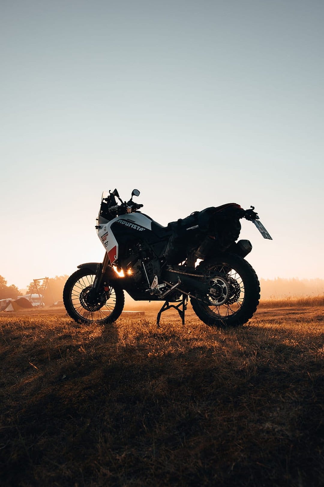 Adventure motorcycle with camping gear parked on grass during a hazy golden hour sunrise at a remote campsite.