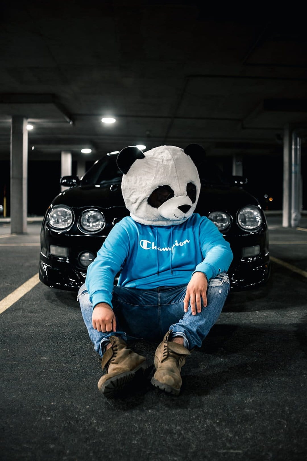 Person in a panda mask and blue hoodie sits on the ground in a garage beside a black car in this urban scene.