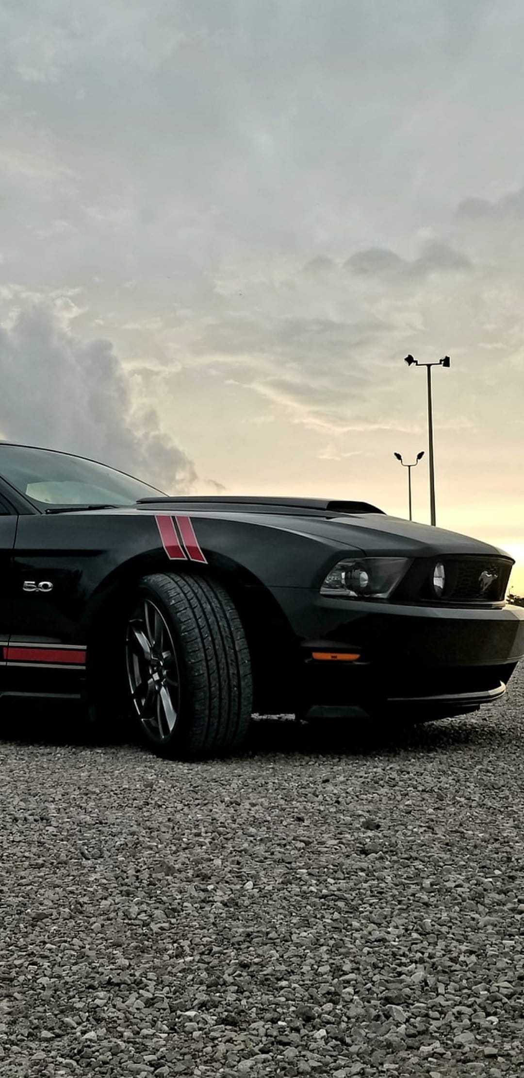 Black Mustang GT 5.0 with red stripes parked on a gravel lot under a cloudy sunset sky in a low-angle view.