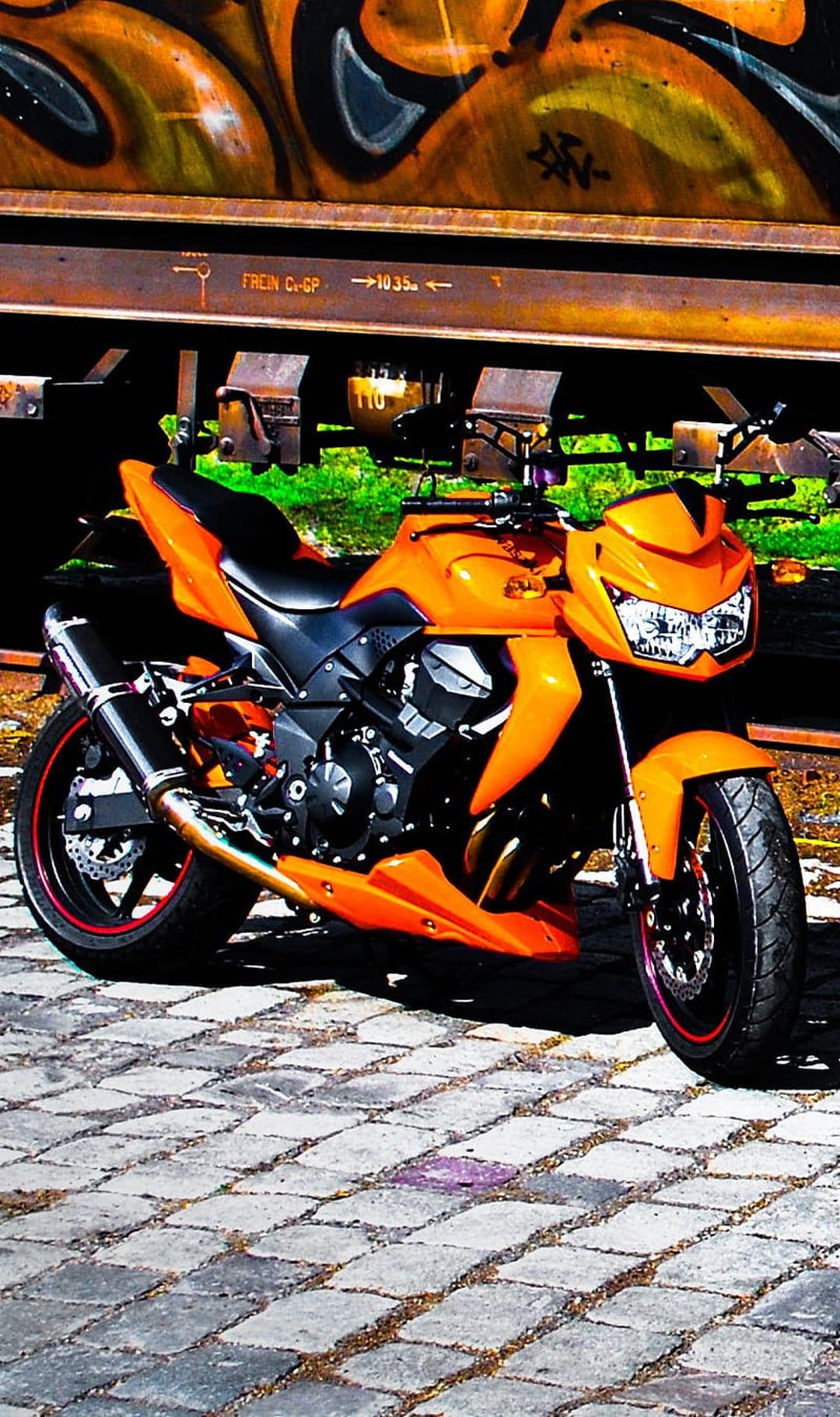 Orange sport motorcycle parked on cobblestones in front of a vibrant graffiti-covered industrial train car.
