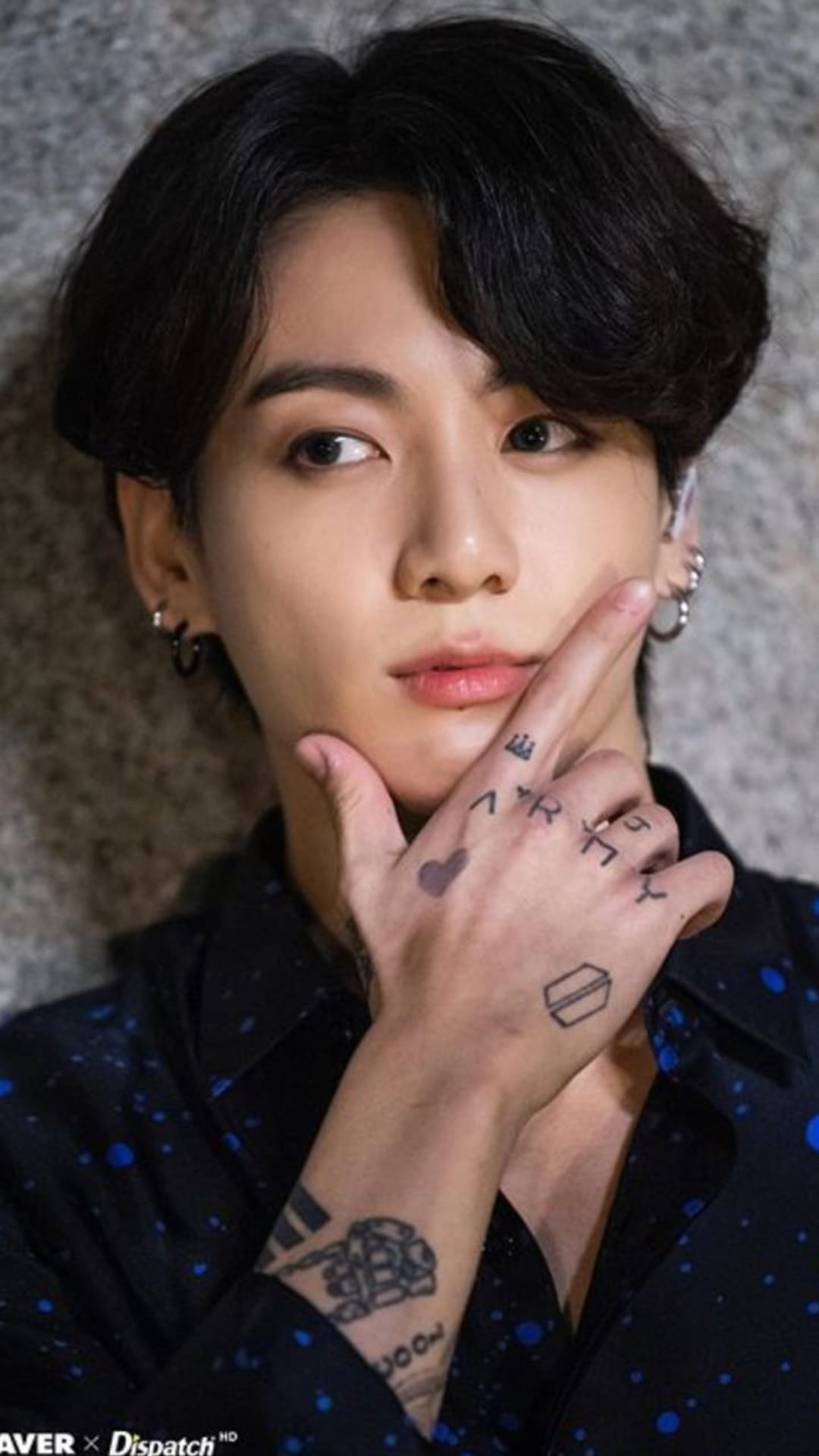 Jungkook rests a hand with ARMY tattoos on his chin while wearing silver earrings and a blue-speckled shirt.