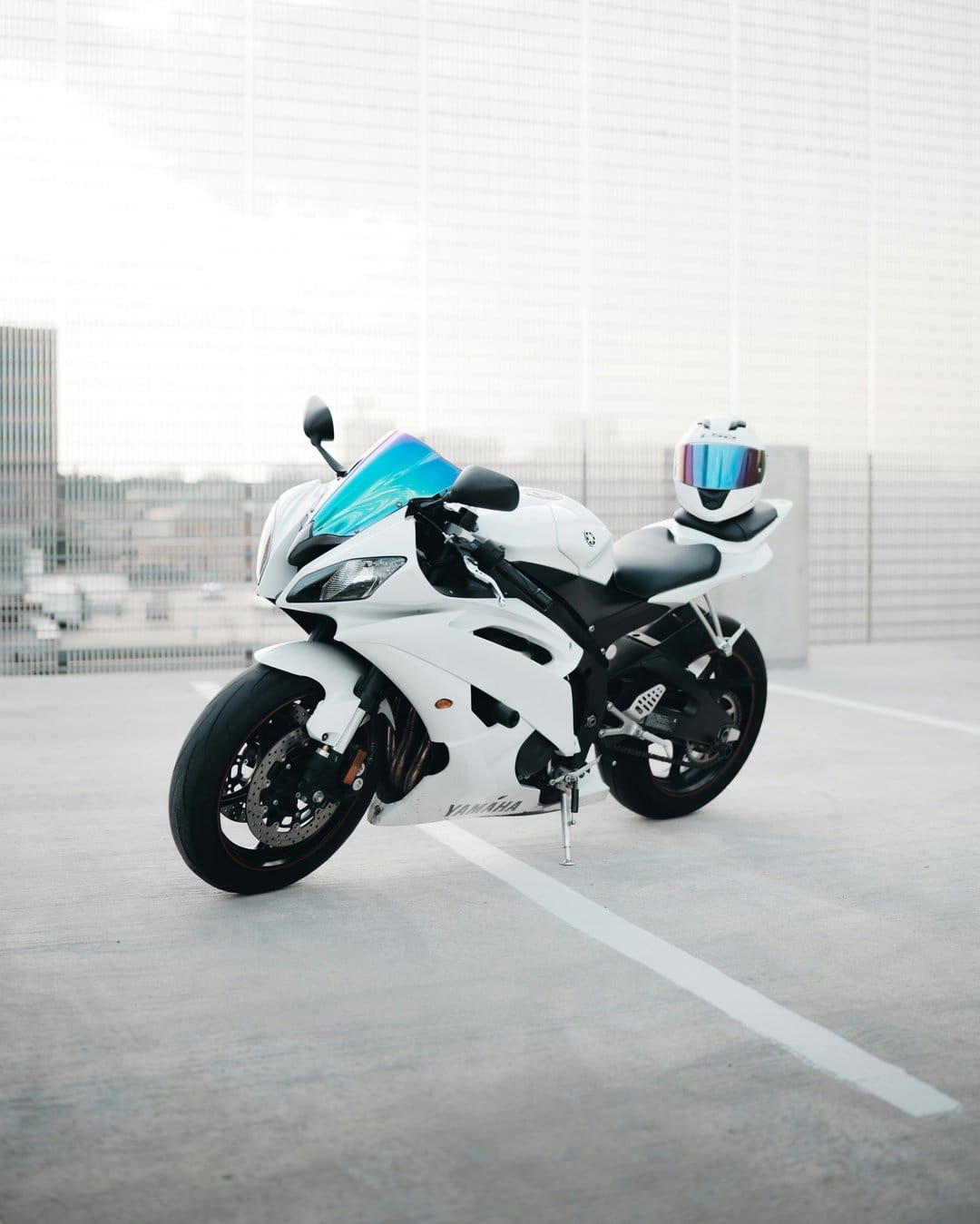 White Yamaha motorcycle with a blue windshield and matching helmet parked against a blurred city skyline backdrop.