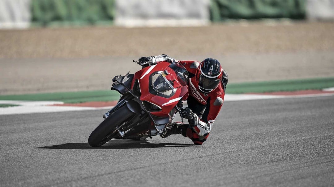 Red Ducati superbike leaning deep into a racetrack corner with a rider in matching professional racing leathers.