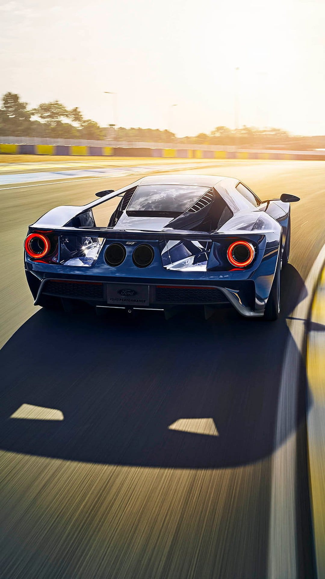 Ford GT supercar with glowing circular taillights racing on a track during a vibrant golden hour sunset.