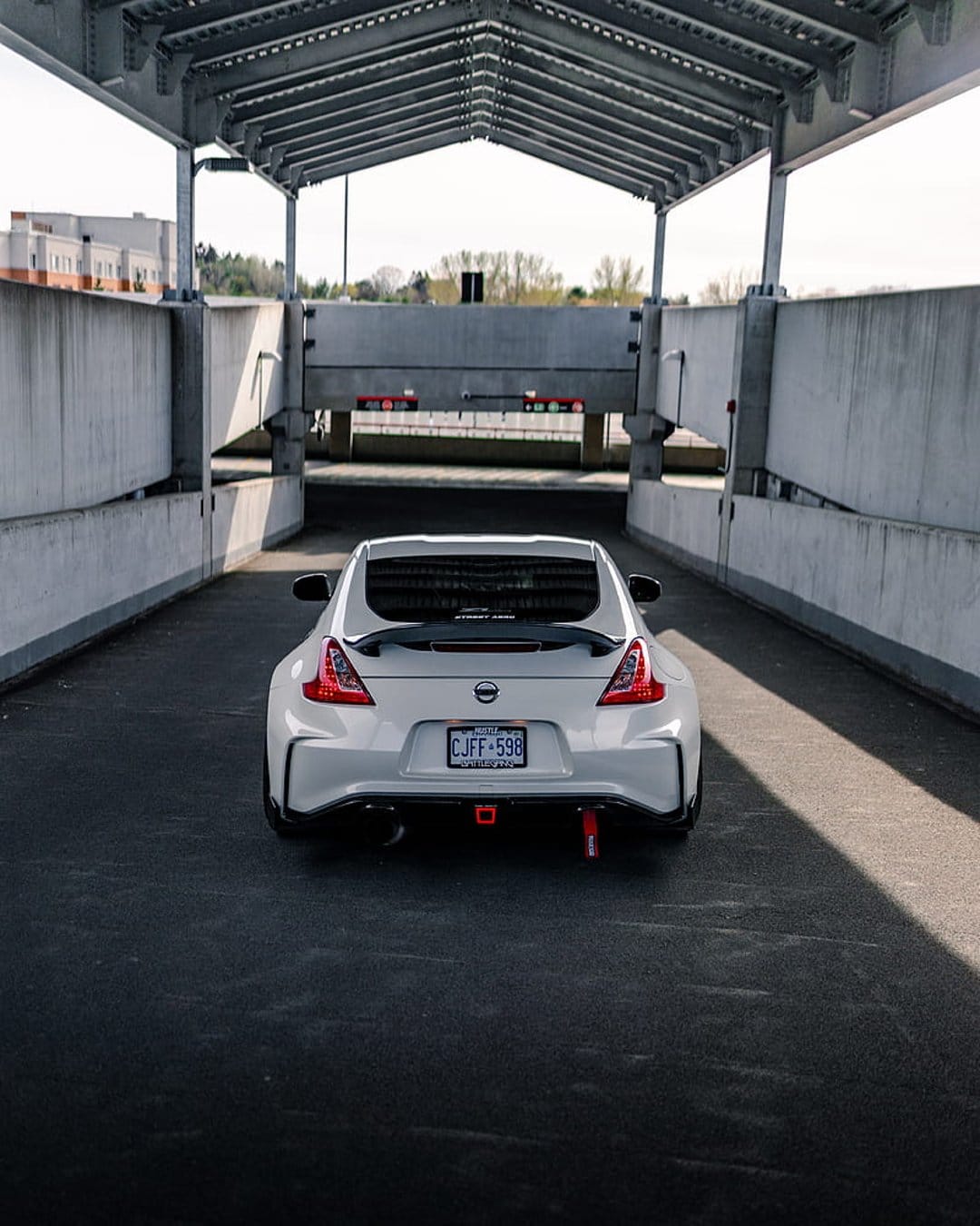 Nissan 370Z rear bumper with red tow hooks and spoiler in a concrete underpass with dramatic sunlit shadows.