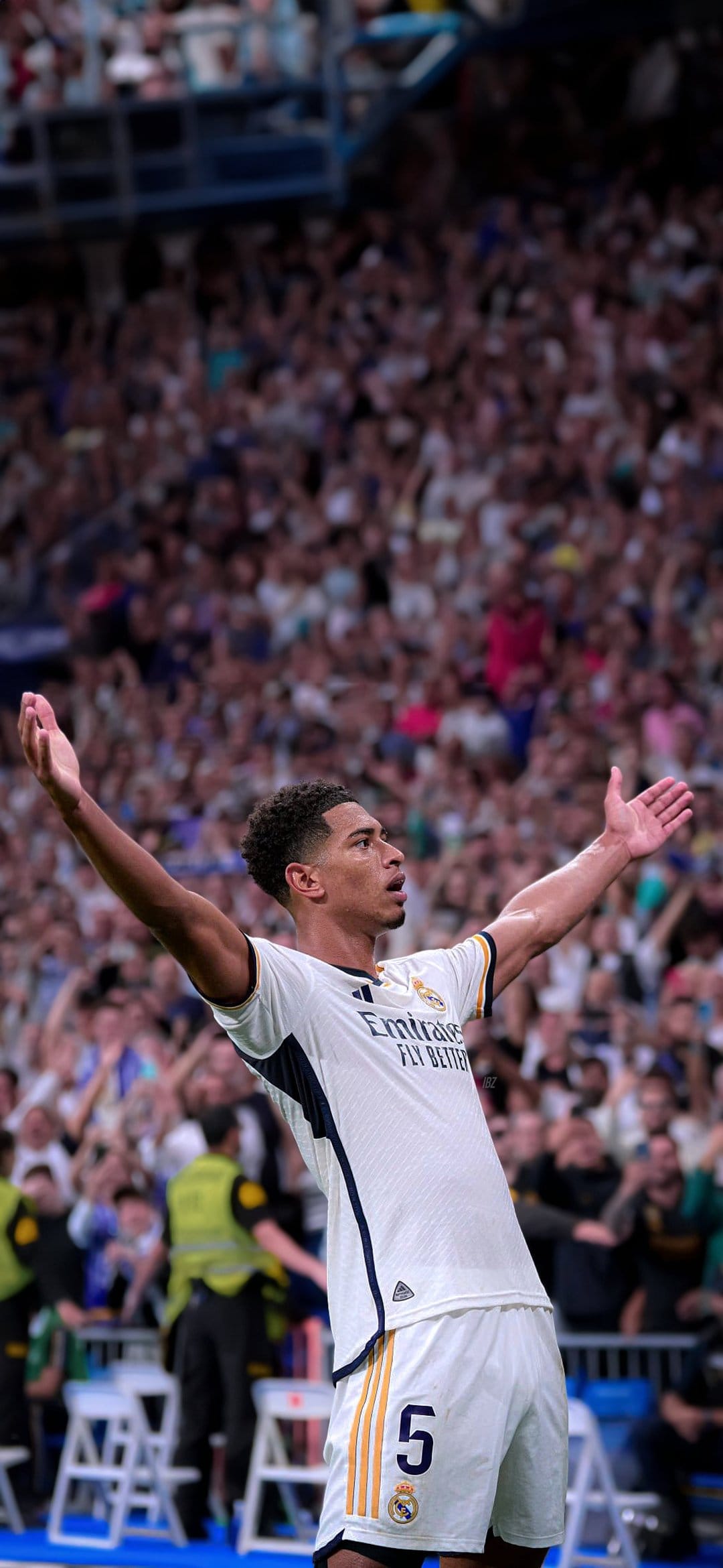 Jude Bellingham raises his arms in celebration while wearing a white Real Madrid kit in a crowded soccer stadium.