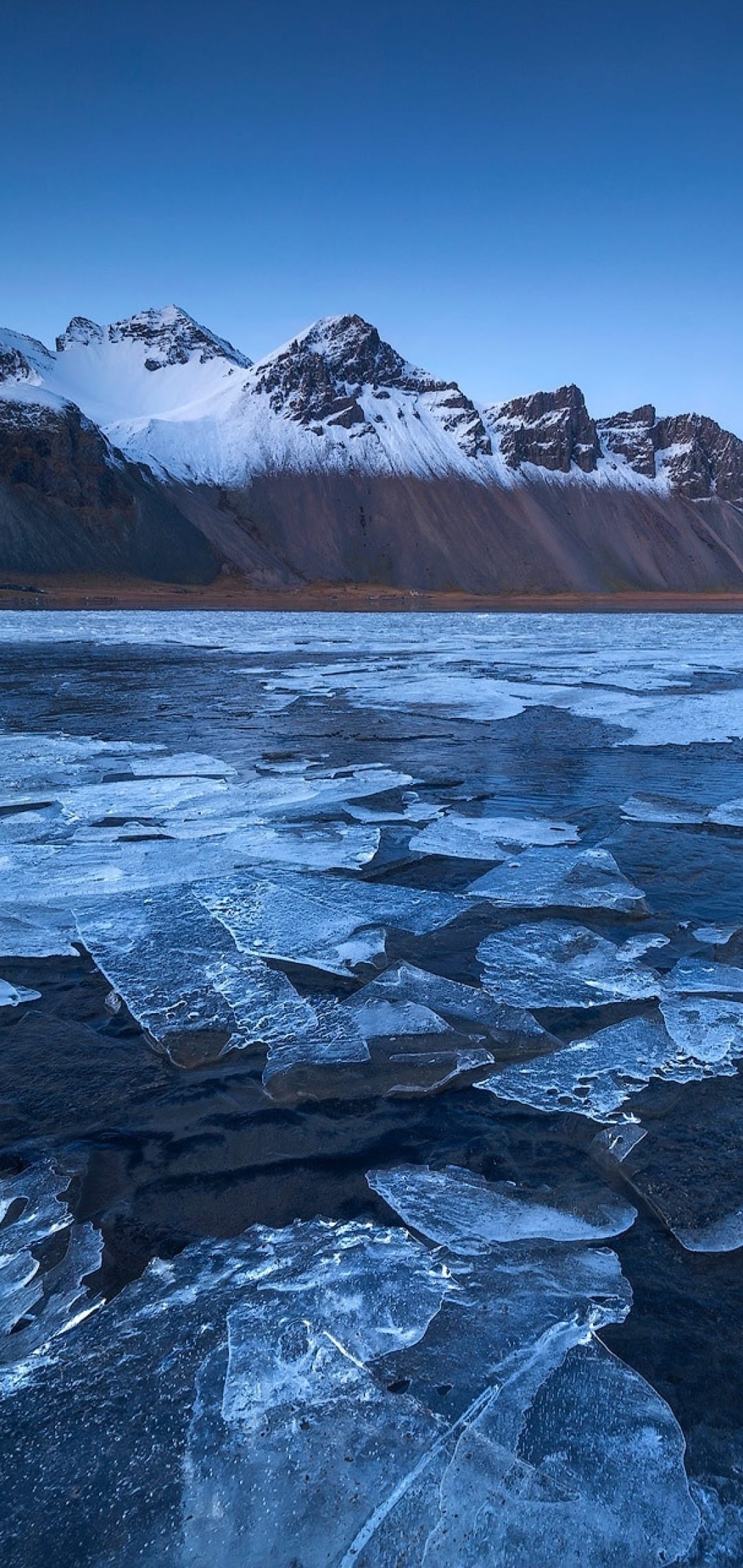 Crystalline ice shards on a frozen shoreline leading to snow-capped mountains under a clear blue arctic sky.