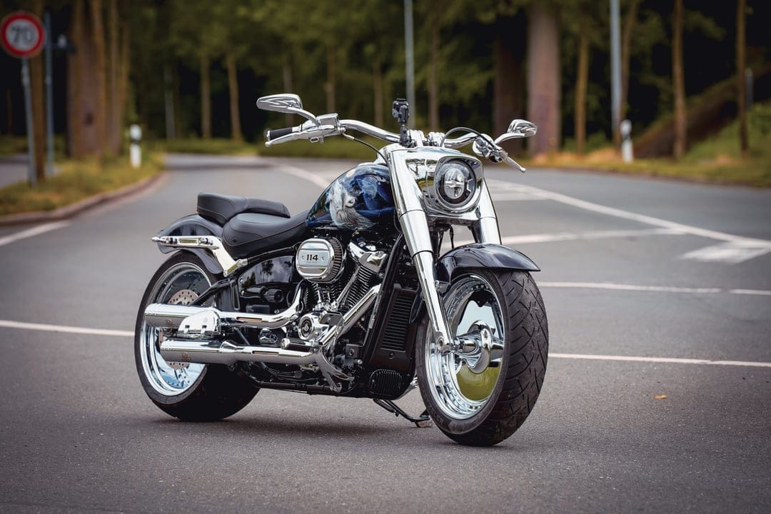 Custom blue Harley-Davidson Fat Boy with chrome accents and solid disc wheels parked on a forest road.