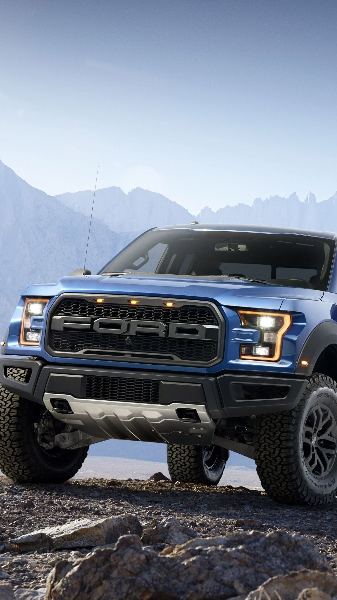 Blue Ford Raptor pickup truck parked on rocky desert ground with mountains under a clear blue sky.