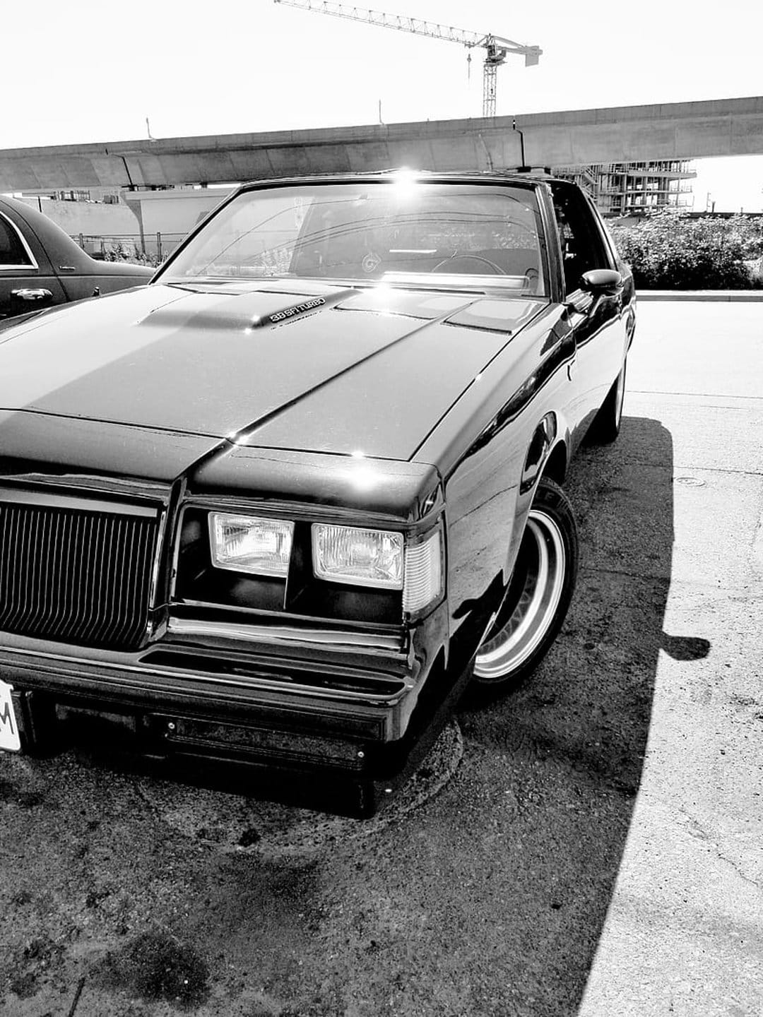Black Buick Grand National muscle car parked on asphalt under a concrete overpass and industrial crane in sunlight.