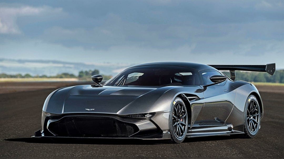 Dark grey Aston Martin Vulcan hypercar on an asphalt runway under a cloudy sky with a large rear wing.