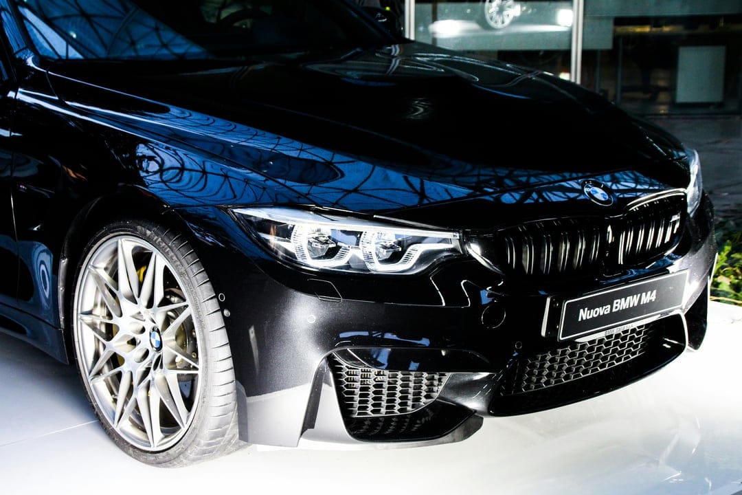 Black BMW M4 front bumper and headlights reflecting a bright environment with detailed alloy wheel spokes.