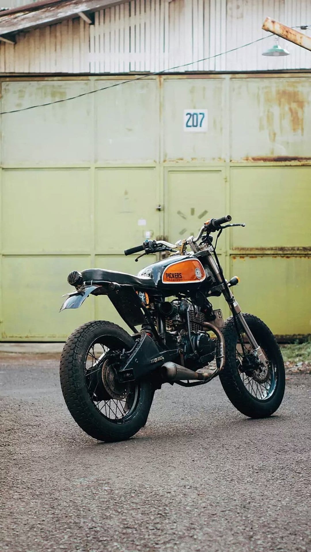 Custom black and orange scrambler motorcycle parked on asphalt in front of an olive green garage with number 207.