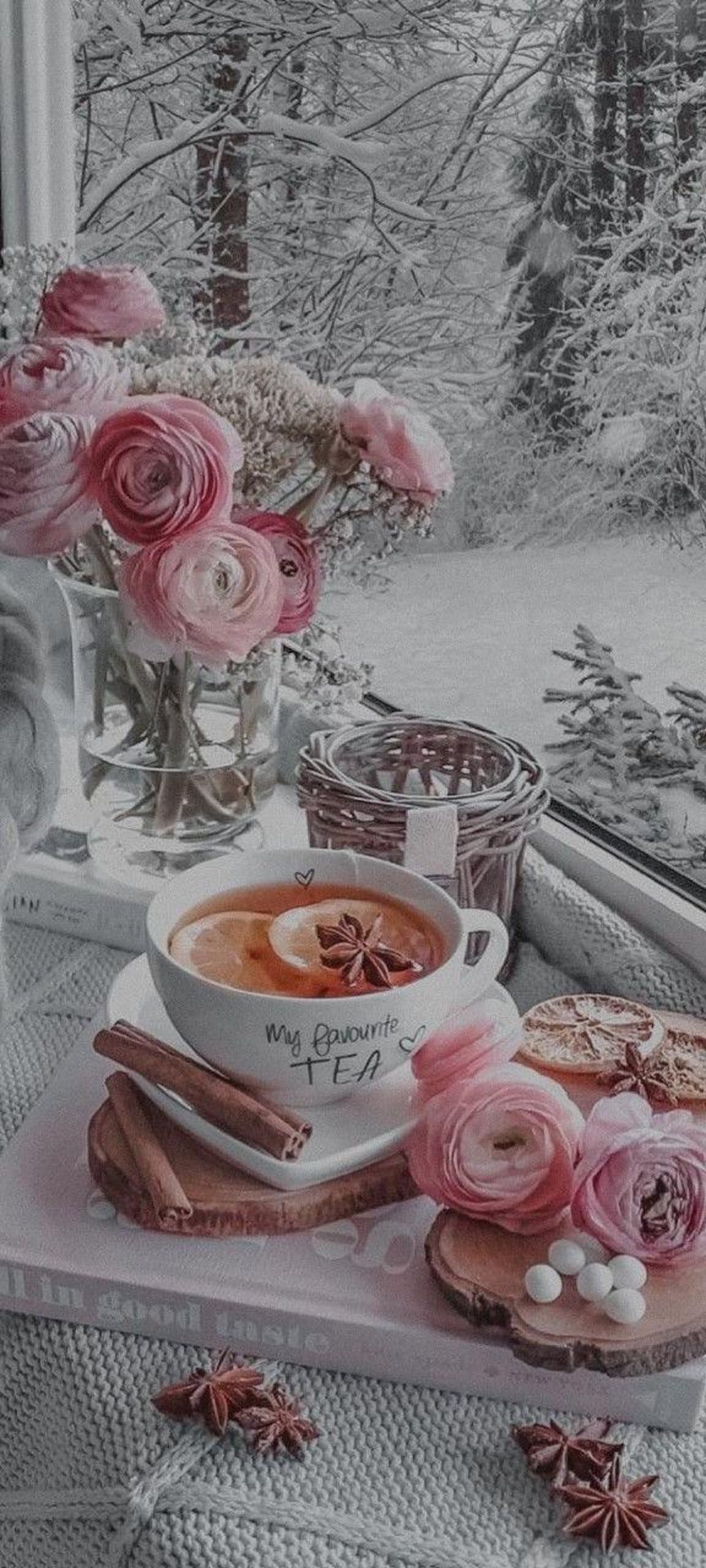 Steaming tea cup with lemon slices and cinnamon sticks beside pink ranunculus flowers near a snowy window view.