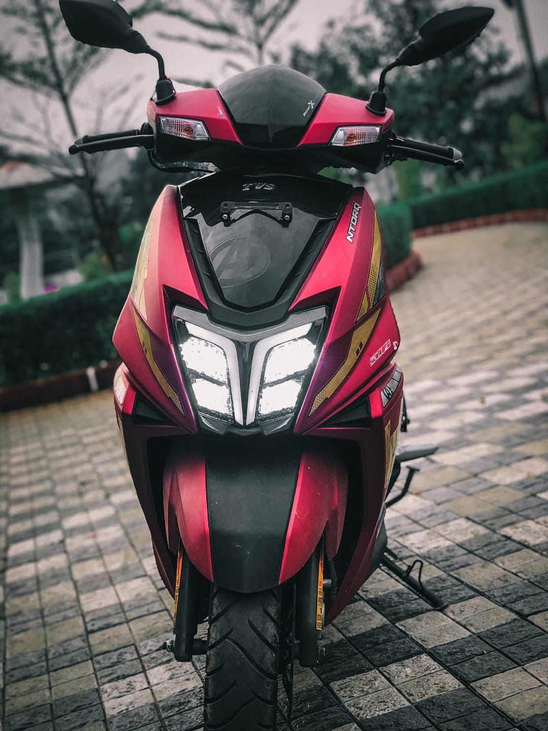 Red and gold TVS NTORQ 125 scooter with glowing LED headlights against a soft green garden background.