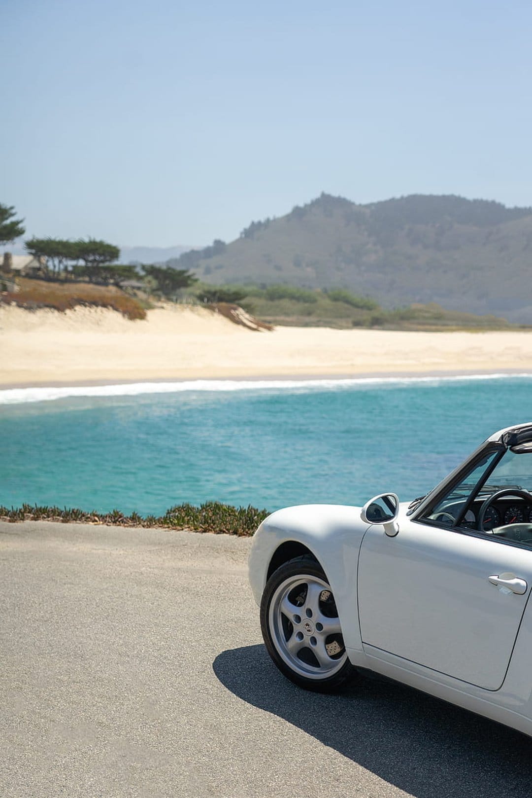 White Porsche convertible parked on a coastal road overlooking turquoise ocean waves and green hills under a blue sky.