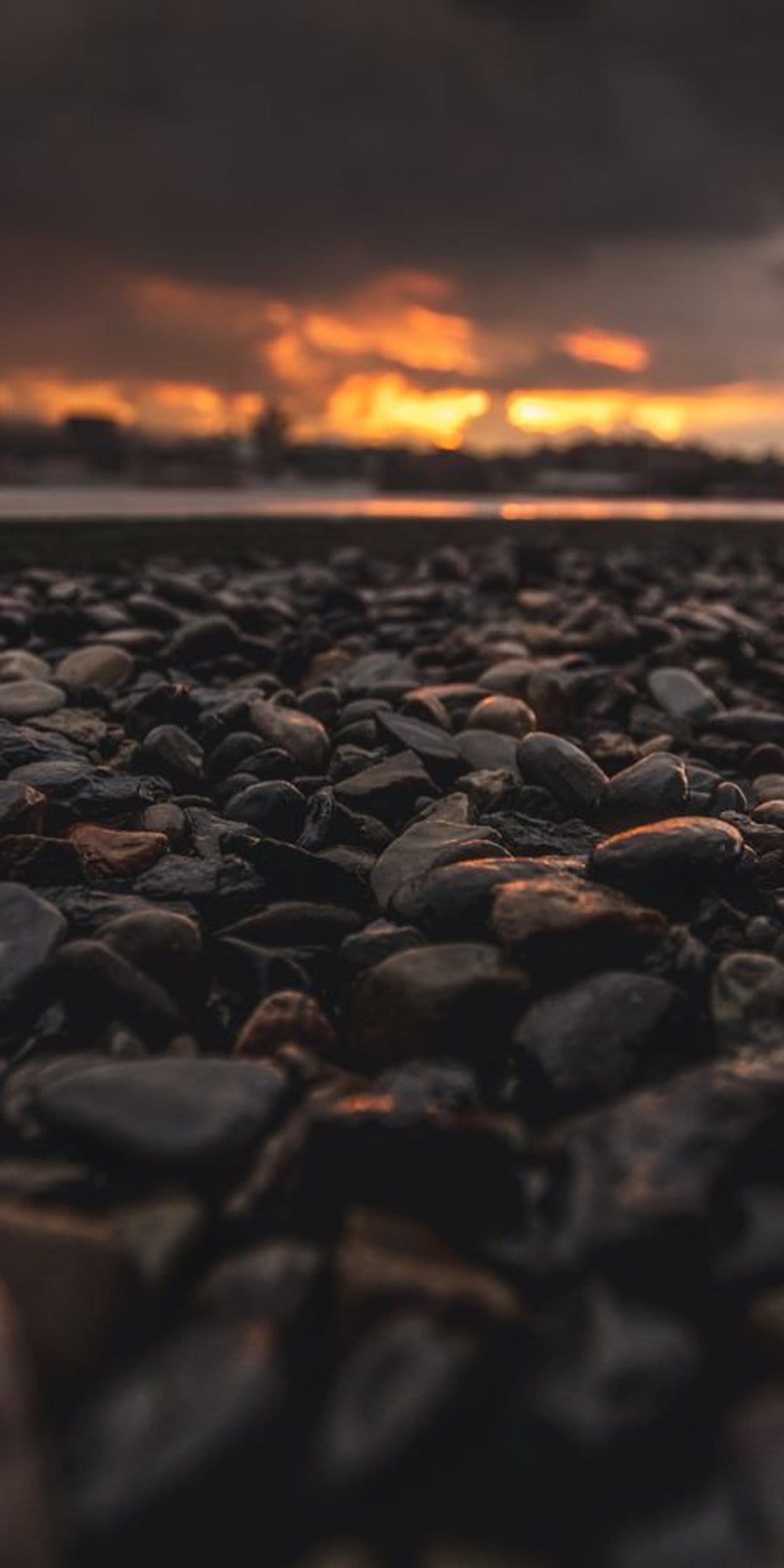 Smooth dark pebbles in the foreground lead to a bright orange sunset over a distant coastal ocean horizon.