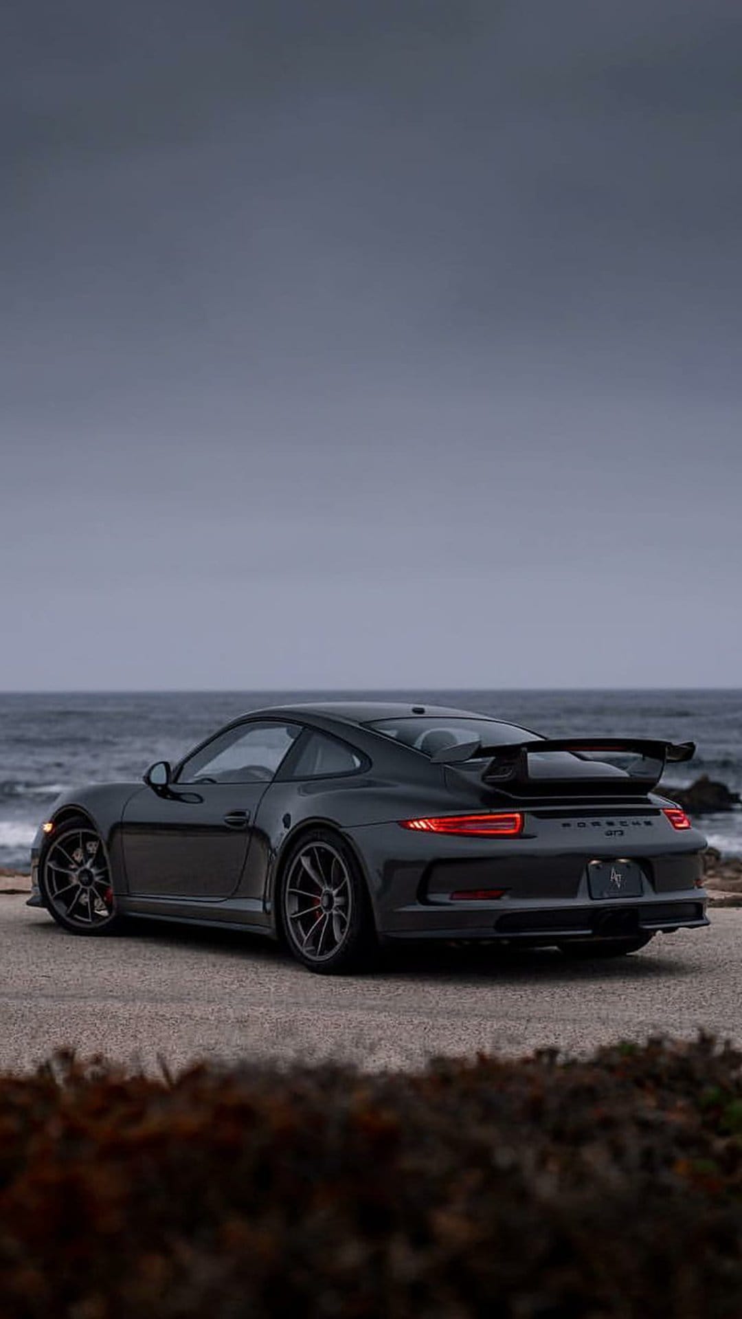 Porsche 911 GT3 with a large rear wing and red calipers parked on an asphalt road overlooking the ocean shore.