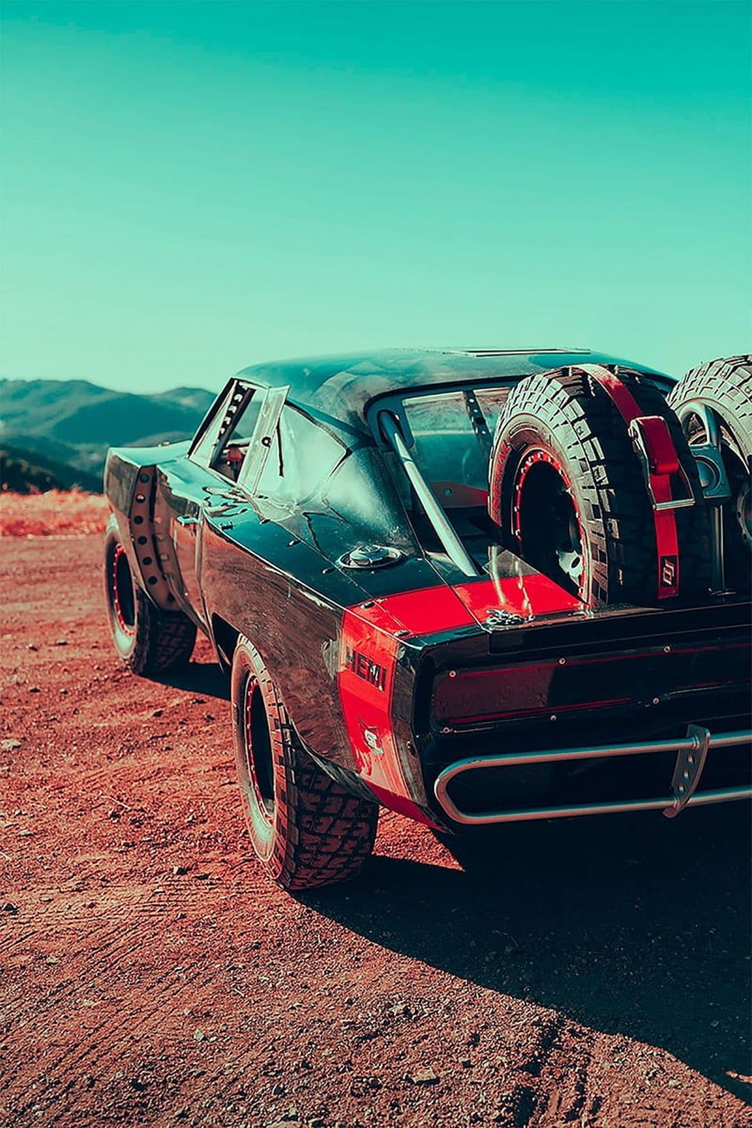 Black Dodge Charger with off-road tires and rear-mounted spares sits on a dusty desert under a blue sky.