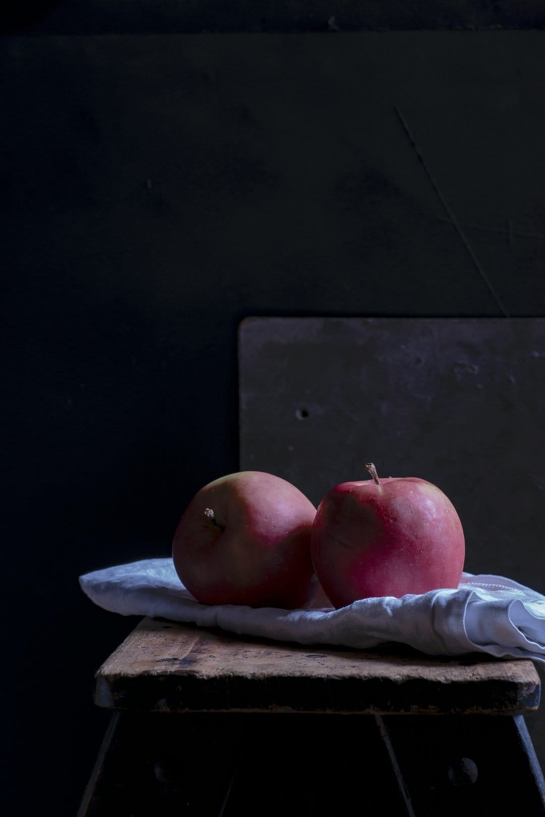 Red apples rest on a white cloth and rustic wooden stool against a dark, textured, moody background.