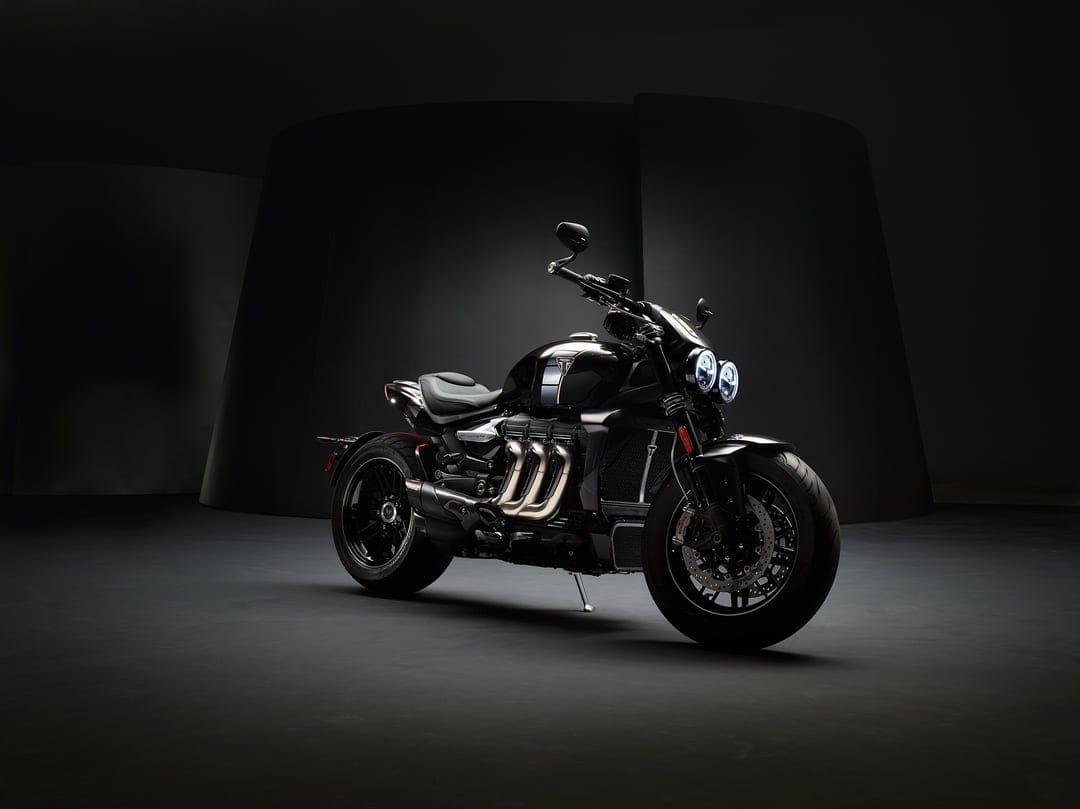 Black Triumph Rocket 3 motorcycle with glowing headlights and triple exhaust pipes in a dark studio setting.