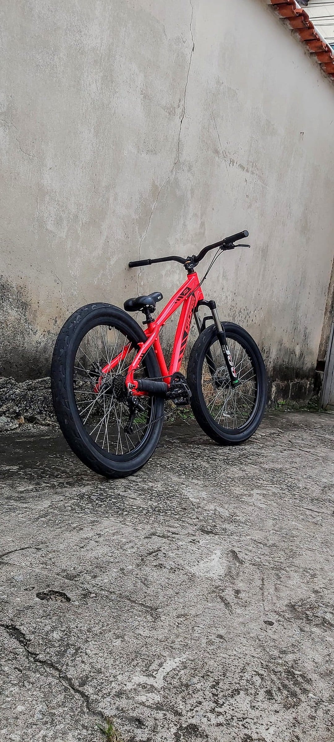 Red mountain bike leans against a gray textured wall with black tires and handlebars on a paved urban ground.