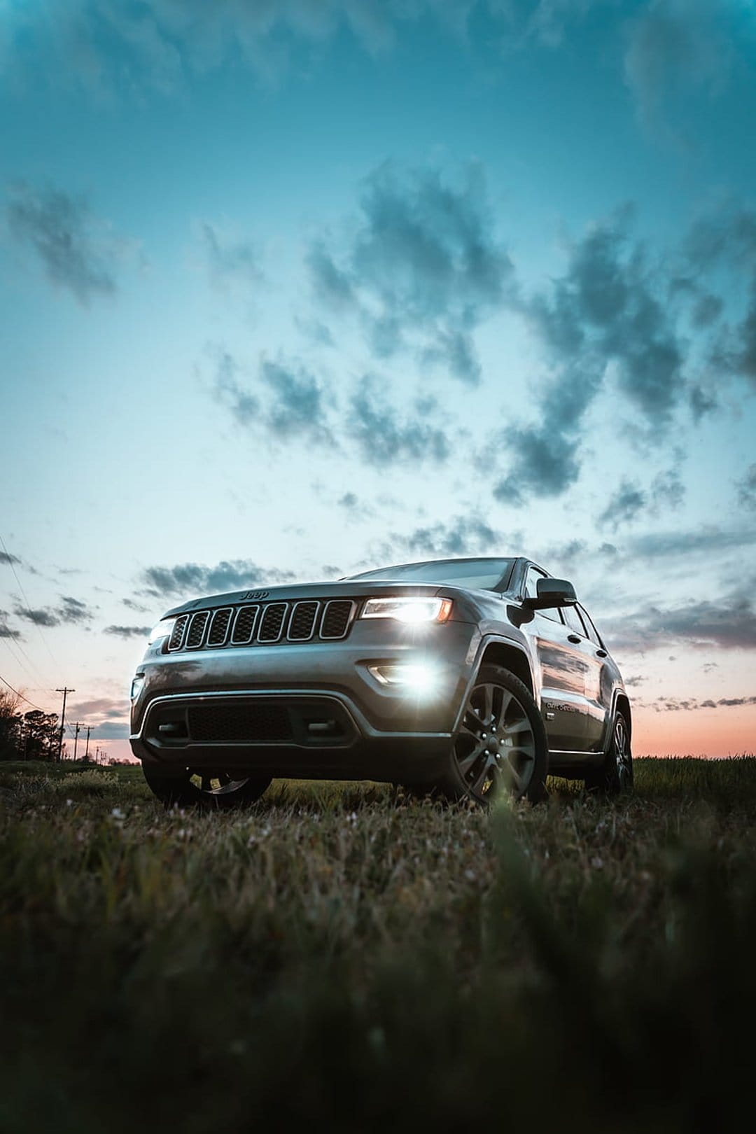 Jeep Grand Cherokee with lit headlights parked in a grassy meadow under a dramatic orange and blue sunset sky.