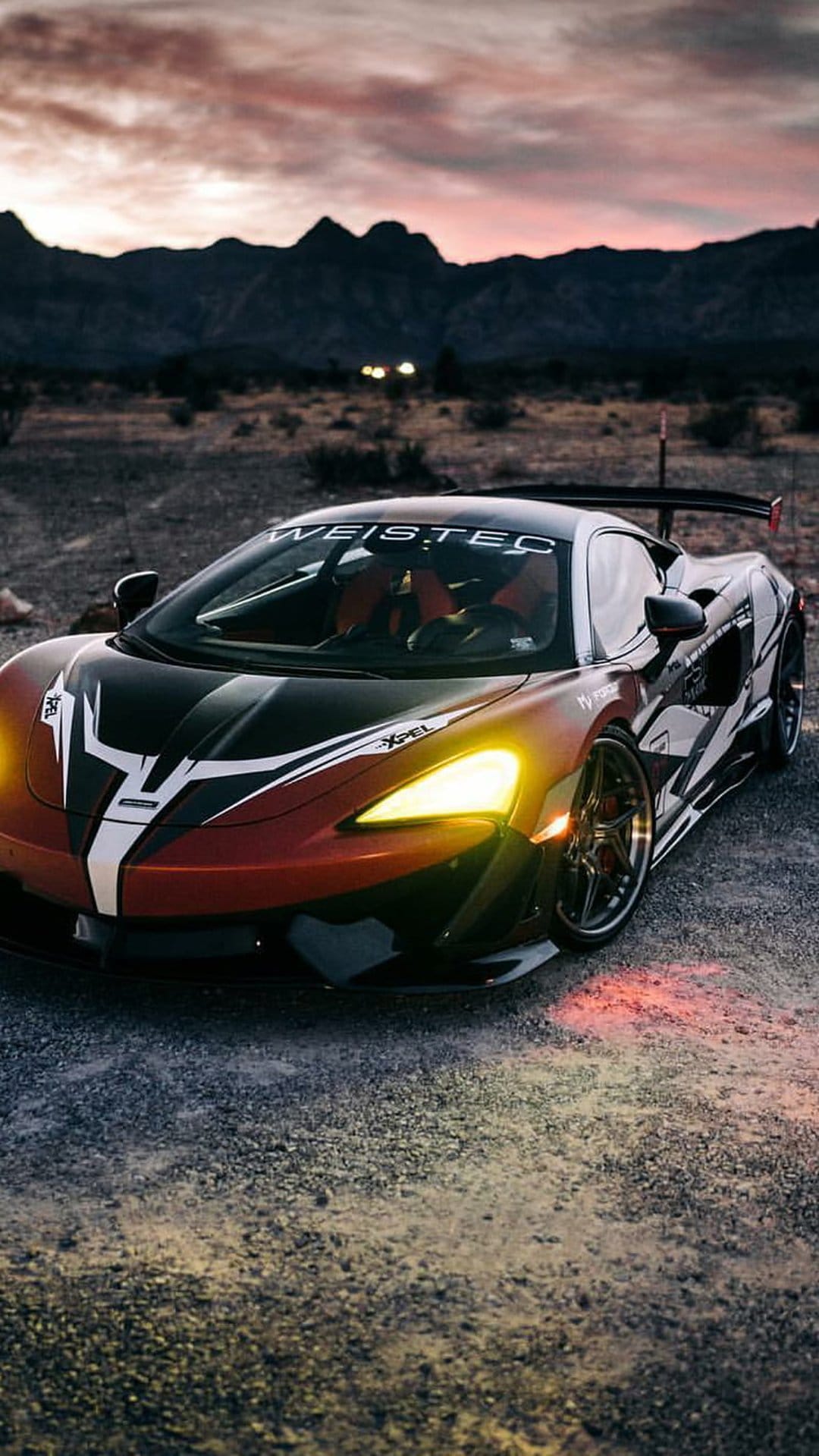 McLaren supercar with red accents and glowing headlights parked on a desert gravel road during a purple sunset.