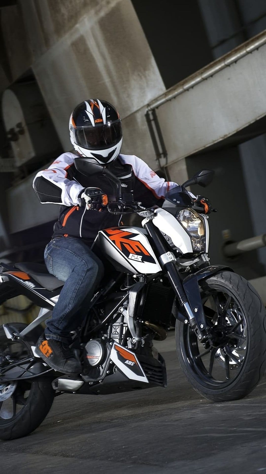 KTM 125 Duke rider in orange and white racing gear leans into a turn beneath an industrial concrete structure.