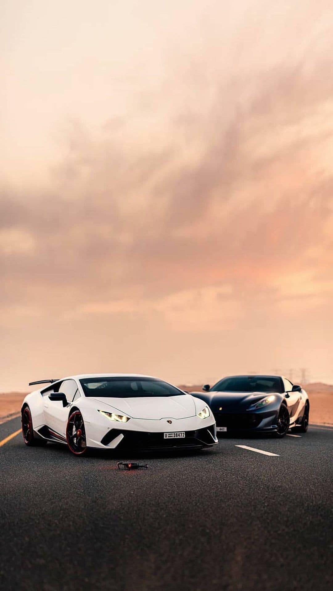 White Lamborghini and dark Ferrari parked on a desert asphalt road during a golden sunset with mountain peaks.