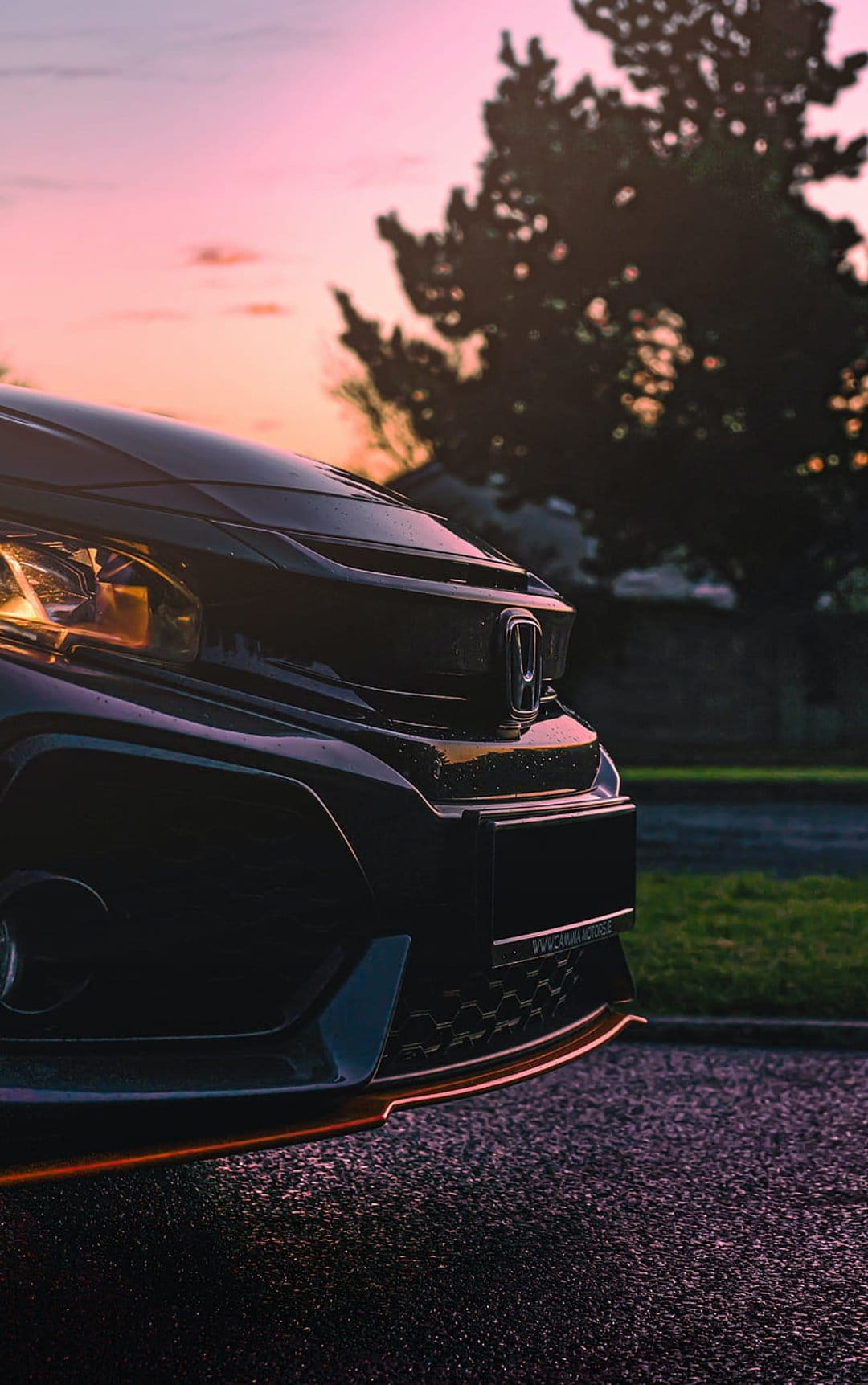 Dark sports car front with orange trim and headlights under a pink sunset sky with a silhouetted tree.