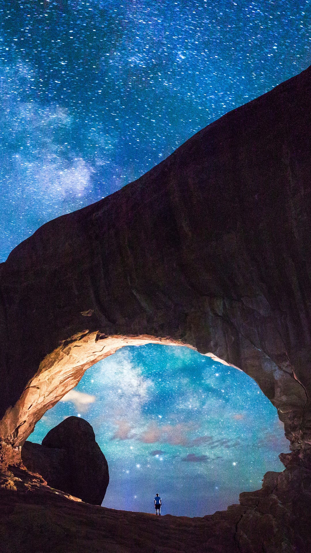 Person standing under a large rock arch at night looking up at the glowing Milky Way galaxy in a starry sky.