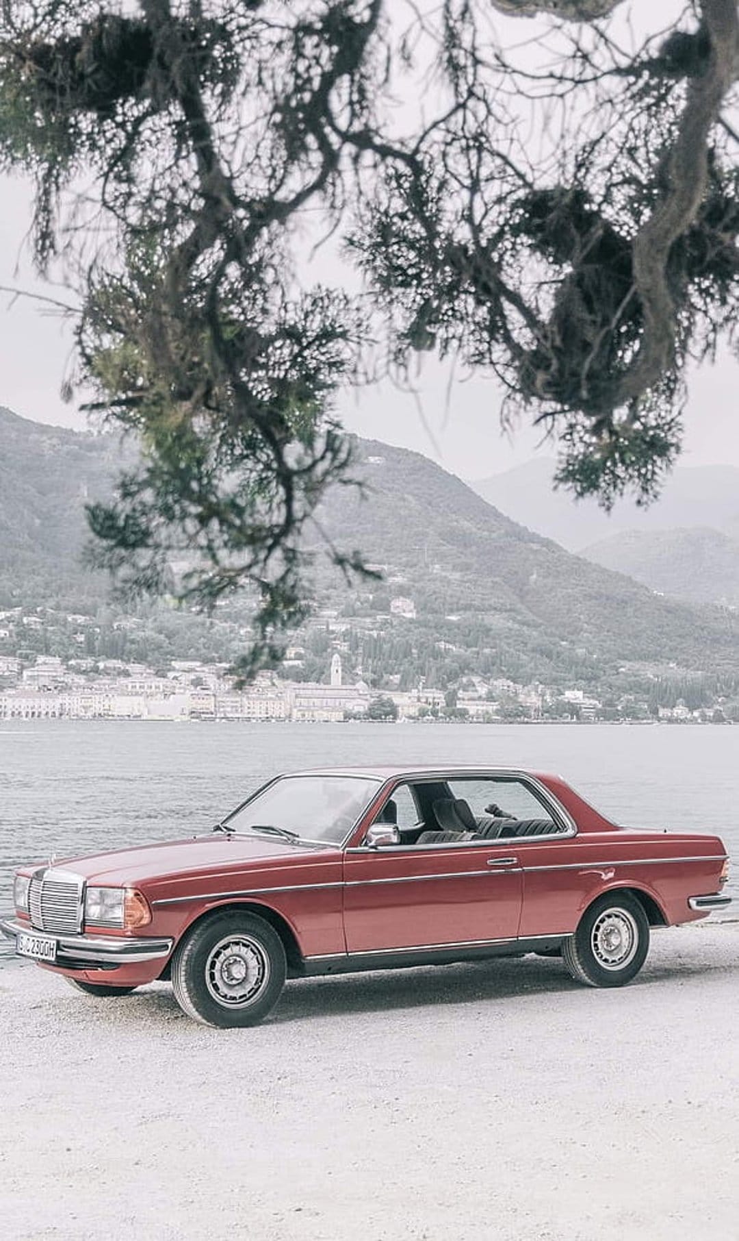 Maroon Mercedes-Benz coupe parked on a sandy beach beside a calm lake with mountains and a village behind it.