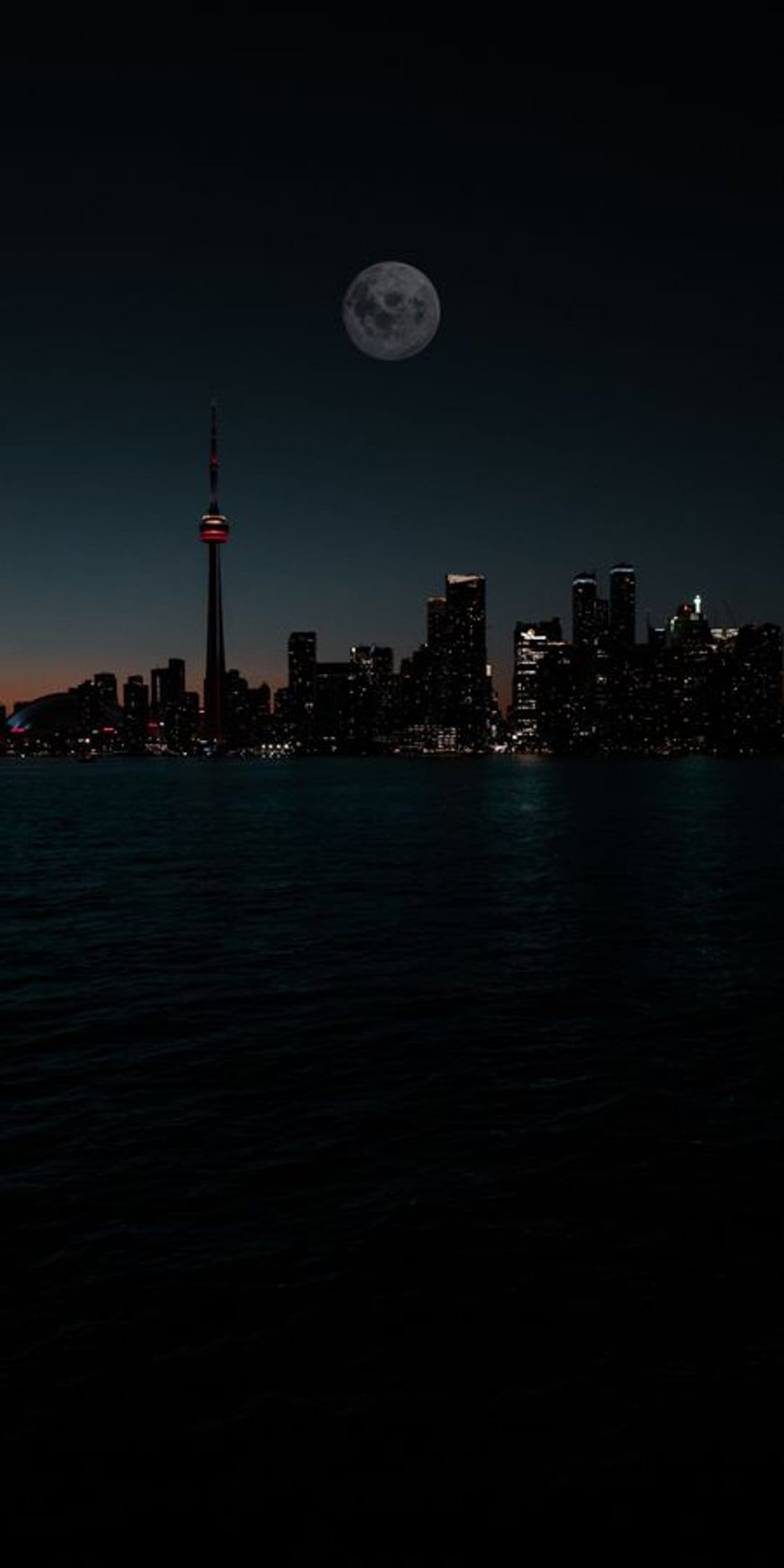 Skyscrapers and a glowing tower stand under a large full moon reflected in the calm harbor water at night.