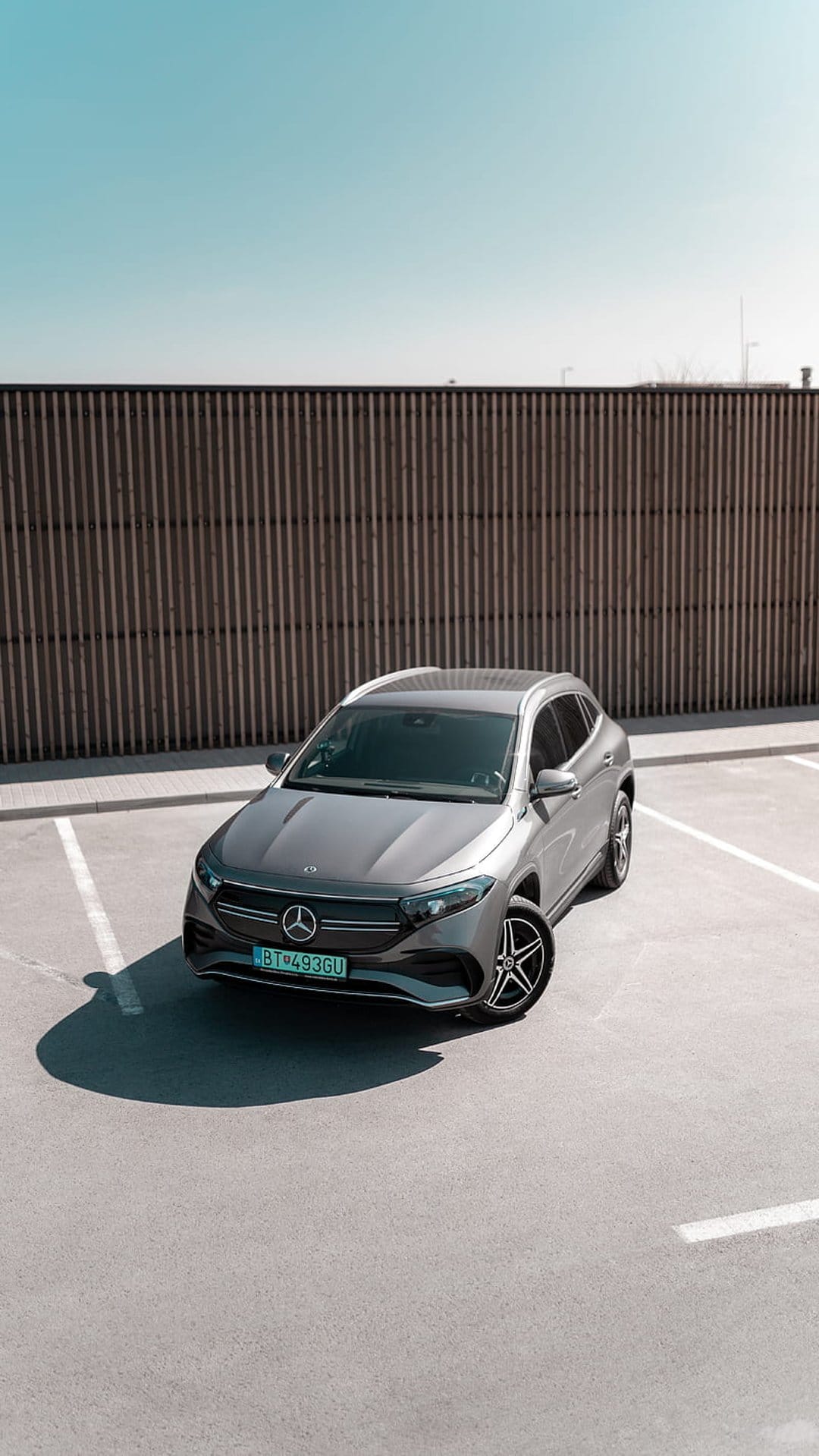 Mercedes EQE SUV in metallic gray parked on asphalt with a wooden fence and blue sky in the bright sunlight.