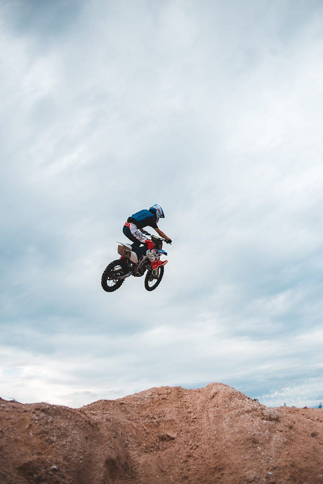 Motocross rider on a red dirt bike performs a high mid-air jump against a backdrop of overcast clouds.
