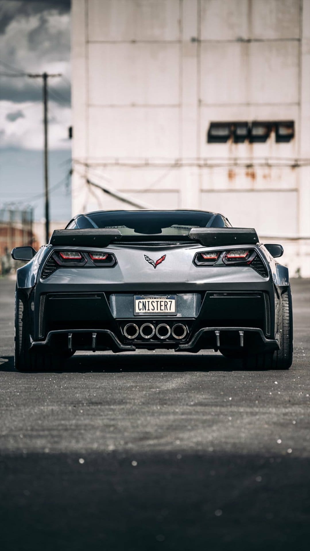 Corvette C7 Z06 rear with a large spoiler and quad exhaust parked on asphalt by a weathered industrial building.