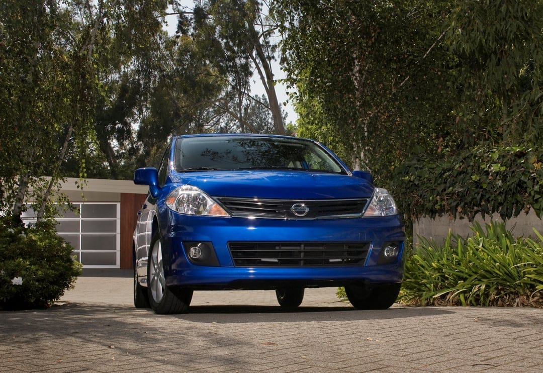 Blue Nissan Versa on a cobblestone driveway framed by lush green trees and a modern garage with frosted glass.