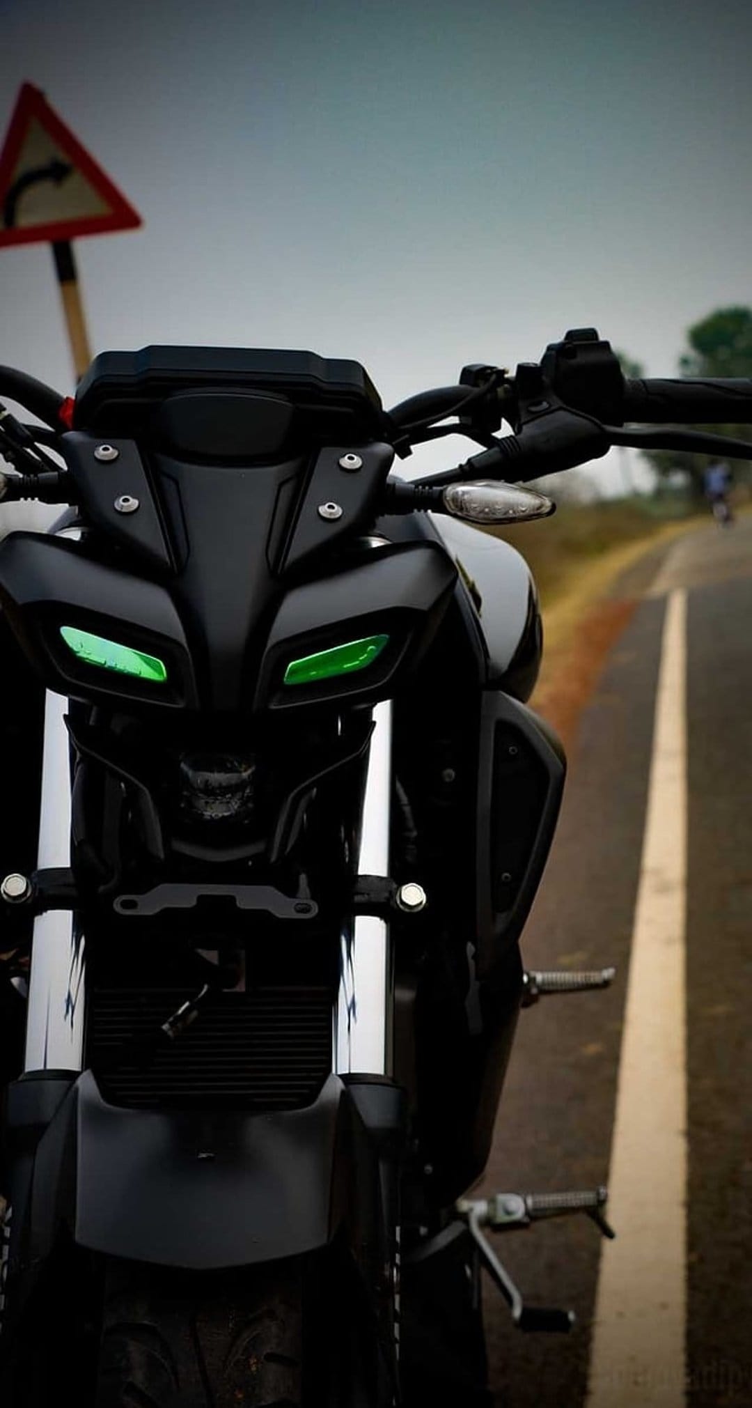Black motorcycle with glowing green headlights on a wet road next to a triangular caution sign under overcast skies.