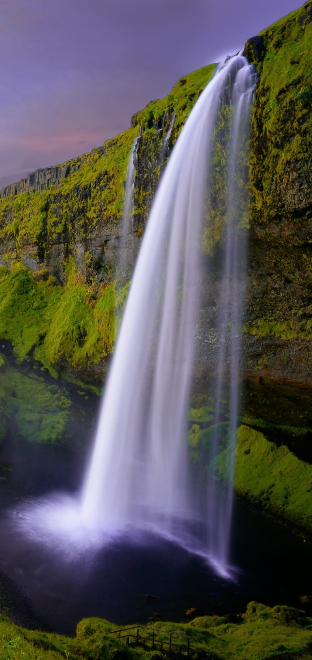Seljalandsfoss waterfall cascades down a green mossy cliff under a pink and purple sunset sky in Iceland.
