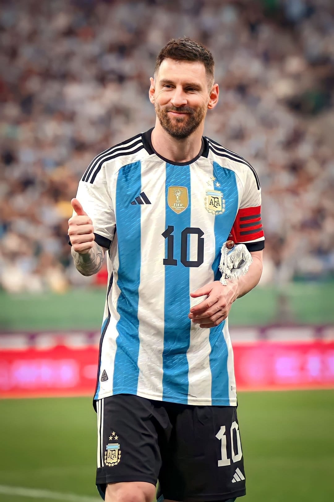 Lionel Messi wearing a blue and white Argentina jersey gives a thumbs up while standing on a football pitch.